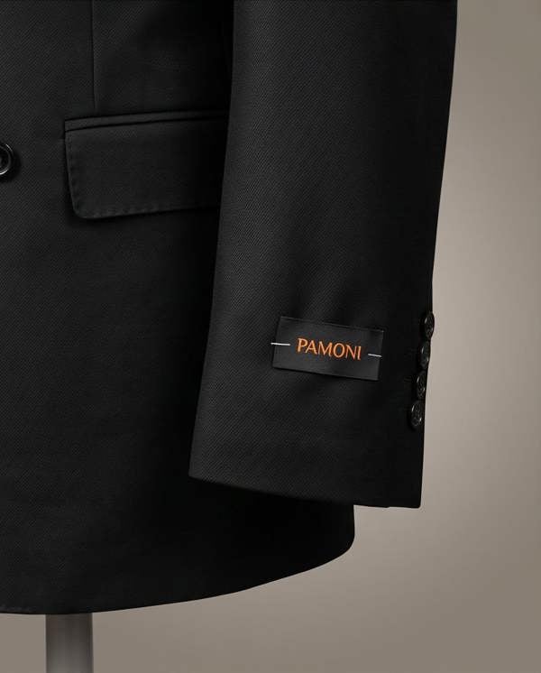 Close-up of a black PAMONI blazer sleeve with a black brand label featuring orange lettering, set against a neutral beige background.