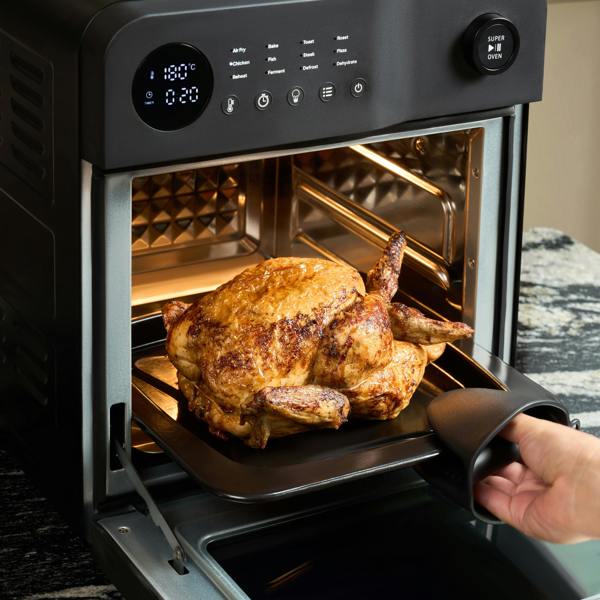 Using an oven tray handle, someone removes a roasted, PFAS-free whole chicken from the Fridja f66 Super Oven - 15L Digital Air Fryer Oven with convection cooking and digital controls, ready to serve.