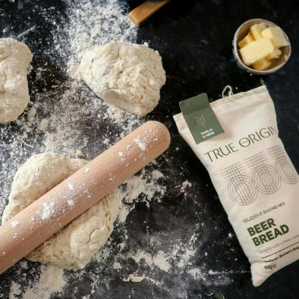 A wooden rolling pin resting across portions of artisanal beer bread dough on a heavily floured black counter, beside a fabric sack of Garlic and Herb mix and a small bowl of cubed butter.