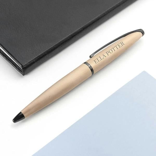 Close-up of a personalised rose gold Cross ATX ballpoint pen engraved with the name 'ELLA POTTER', resting diagonally next to a closed black notebook on a light blue surface