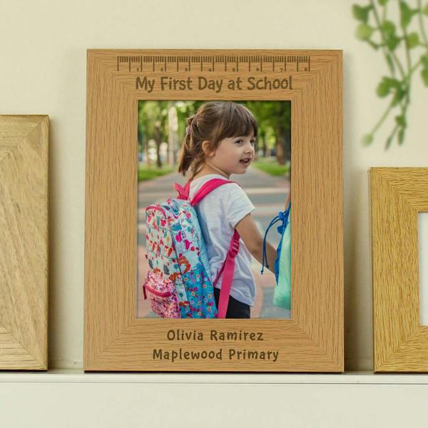 A natural wooden photo frame standing on a shelf. It contains a photograph of a young girl wearing a colourful school backpack. The frame is engraved with a ruler motif, the text "My First Day at School", "Olivia Ramirez", and "Maplewood Primary".