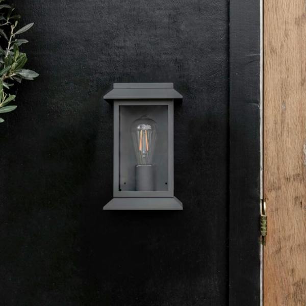 Modern carbon steel lantern wall light with a matte finish and glass framing, mounted on a dark exterior wall next to a natural wood door frame.