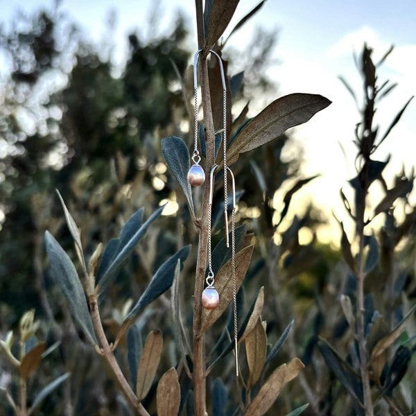 Pair of silver threader earrings with delicate pink pearls, hanging on a branch with leaves.