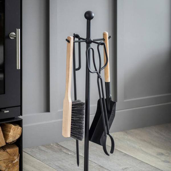 A 4-piece minimalist fireside companion set hanging on a matt black steel stand next to a wood-burning stove filled with logs, showcasing natural beech wood handles, coal scissors, a poker, a shovel, and a brush against a grey panelled wall.