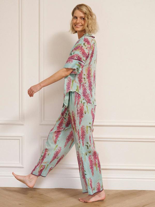 Woman wearing duck egg blue cotton gauze oversized pyjamas with pink wisteria floral print, short-sleeve button-through top and wide-leg trousers, standing barefoot indoors.