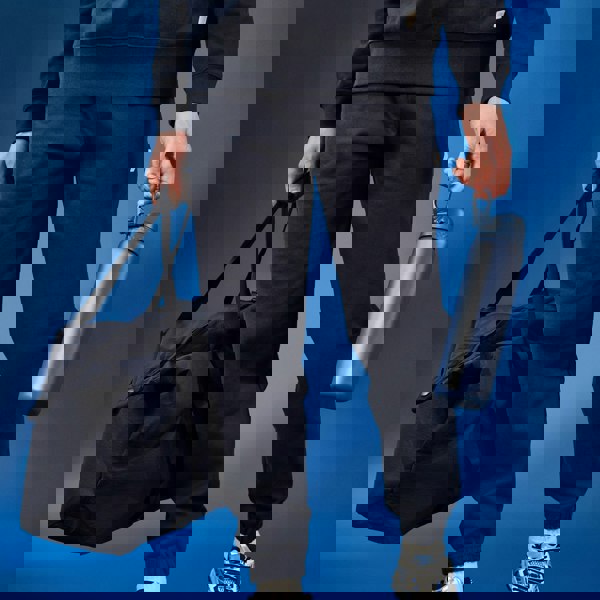 A person carries a dark duffel bag and a dark blue water bottle against a blue backdrop.