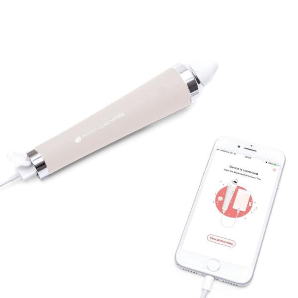 The Rio Pore Perfection, a white electronic handheld device with buttons for personalized skincare and pore cleansing, is shown connected by cable to a smartphone displaying its app interface, both resting on a white surface.