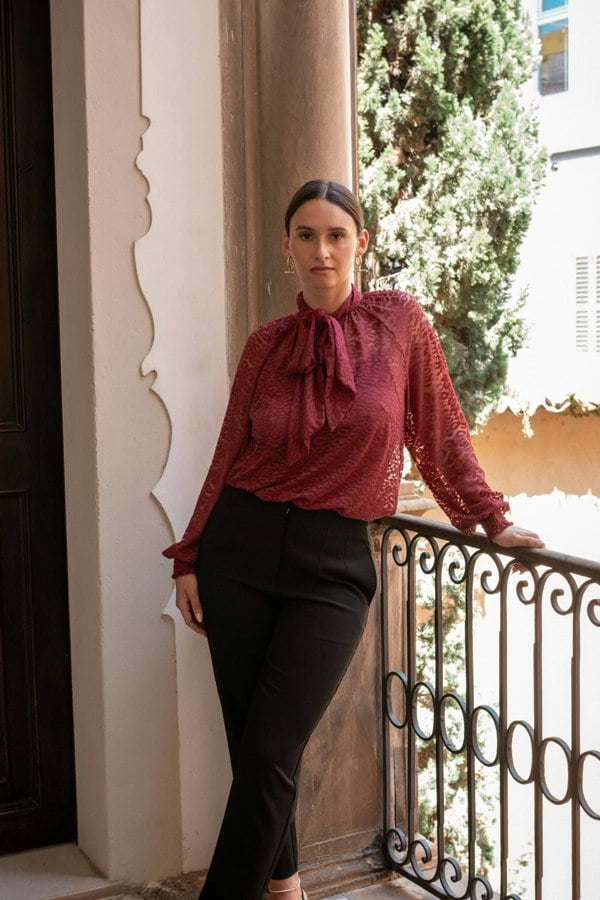 Woman in a red blouse and black pants standing on a balcony with a decorative railing.