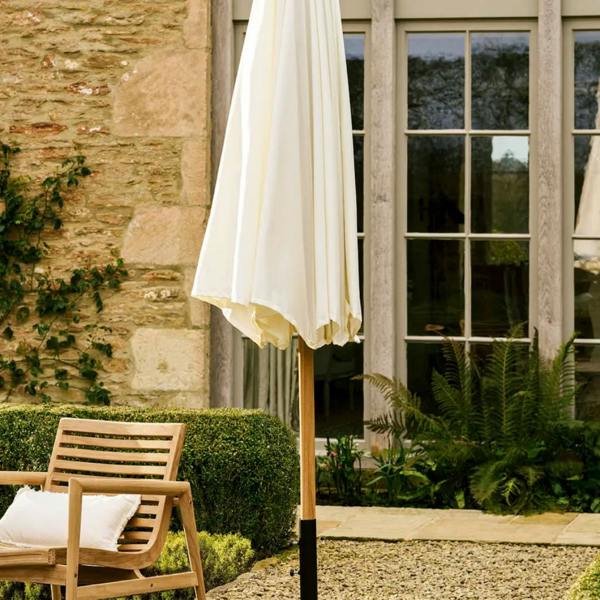A closed and neatly folded 3m natural cream fabric garden parasol on a wooden teak pole standing in a gravel courtyard next to a wooden slatted armchair.