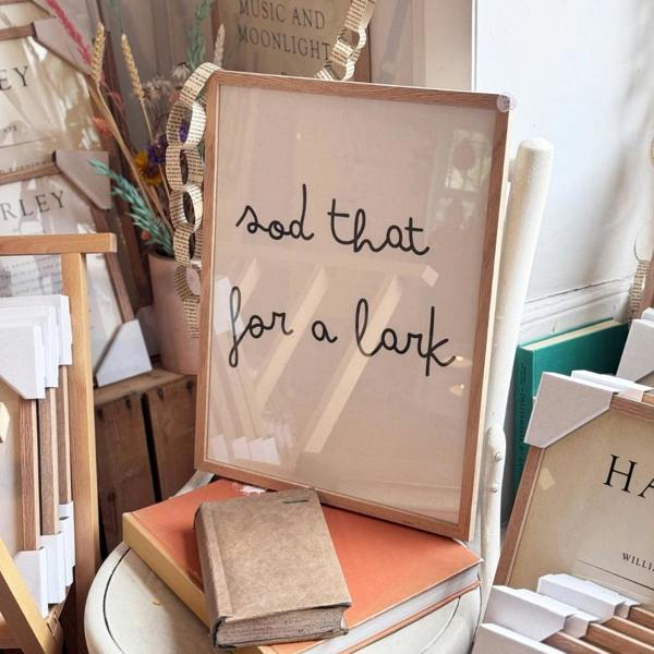 A solid oak framed white cotton typography print displaying the lowercase handwritten-style quote "sod that for a lark" resting on vintage orange books in a shop display.
