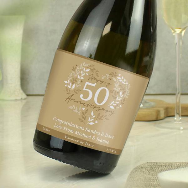 A slightly tilted bottle of personalised 50th Golden Wedding Anniversary Prosecco from Abode Home & Gifts on a marble surface, displaying a gold label with text reading Congratulations Sandra & Dave Love From Michael & Joanne.