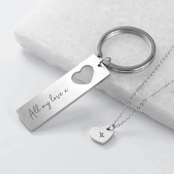 A stainless steel keyring featuring a heart-shaped cutout and the inscription "All my love x", resting next to a matching silver heart pendant necklace engraved with the letter "k" on a textured white surface.