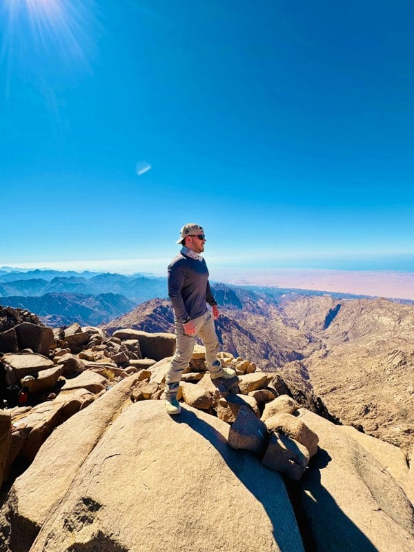 Person standing on a rocky mountain peak with a clear blue sky wearing a Ruffnek hiking snood scarf neck gaiter. Photo copyright Sams_walkabout / Ruffnek 
#HikingGear #HikingAdventures #HikingAccessories #TravelScarf #MountainSnood