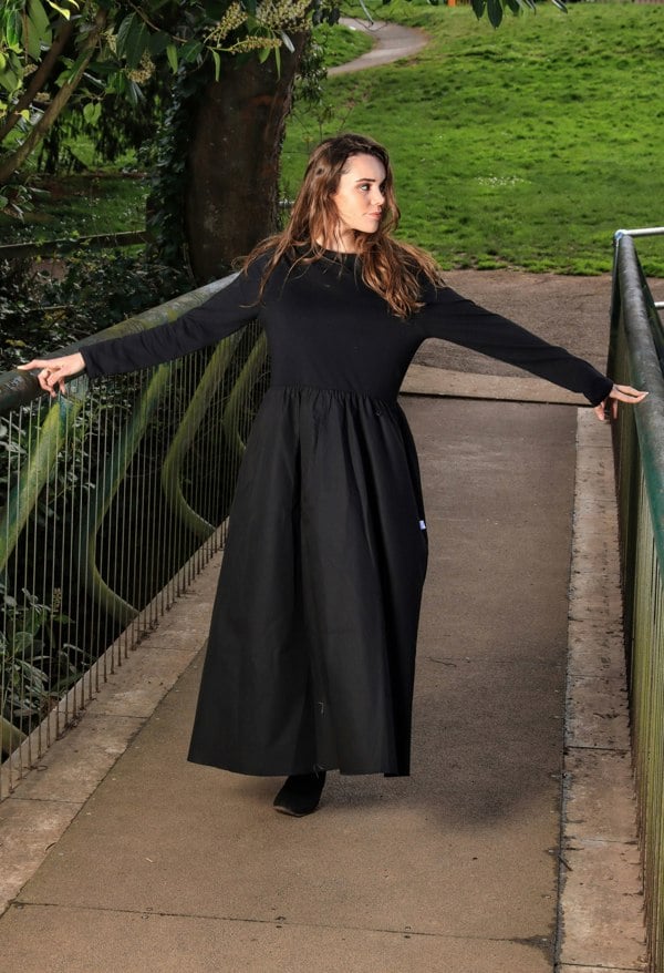 This is a young lady wearing a midi/maxi length black dress with a round neck, long sleeves and a gathered skirt. She is wearing black boots and silhouetted against a woodland background, with her arms outstretched across a bridge.