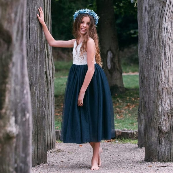 girl in a white top and dark blue skirt standing between two tree trunks in a park.