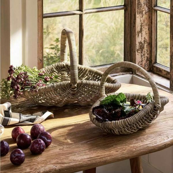 A small and a large hand-woven kubu grey rattan trug basket sitting on a rustic wooden table filled with fresh cut flowers and green salad leaves by a window.