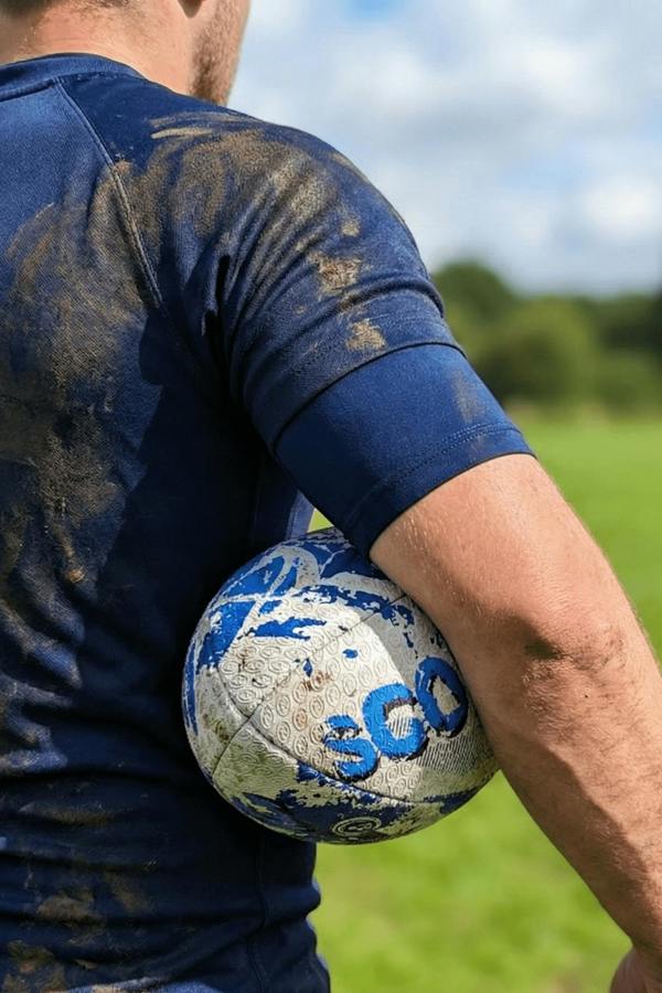 Muddy player holding a blue and white Optimum Scotland rugby ball under his arm on a pitch - Optimum