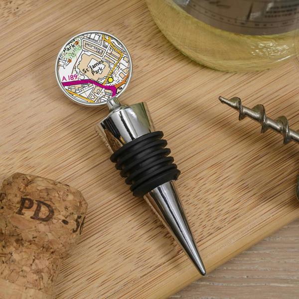 A polished chrome metal Favourite Football Grounds Bottle Stopper featuring a detailed street map of the Emirates Stadium, resting on a wooden board next to a cork.
