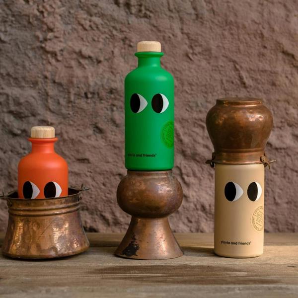 Group display of Yiayia and Friends infused extra virgin olive oil bottles in orange, green, and cream styled with rustic copper kitchenware against a stone wall.