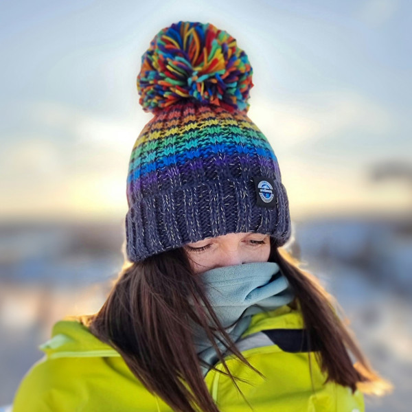 Woman wearing a chunky rib knit bobble hat in rainbow coloured stripes on a navy blue base, The hat also has a deep ribbed headband and contains reflective yarns with a luxury sherpa fleece lining.