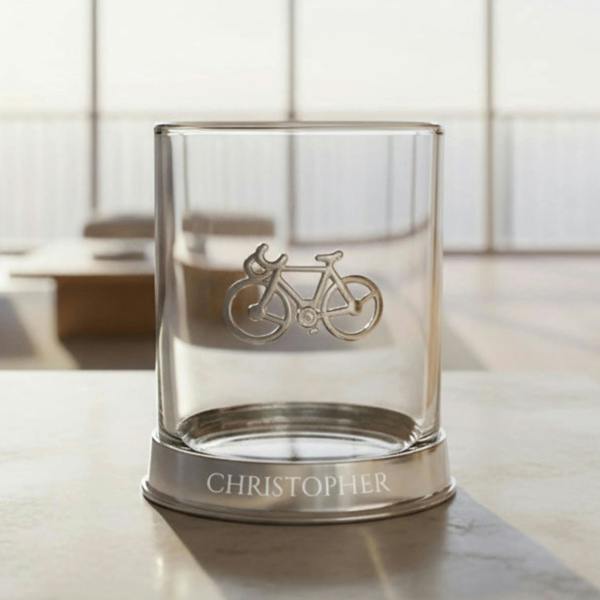 A personalised clear glass whisky tumbler featuring a detailed silver pewter base band embossed with a cyclist motif, custom engraved with the name 'Daddy' on the glass, filled with amber spirit.