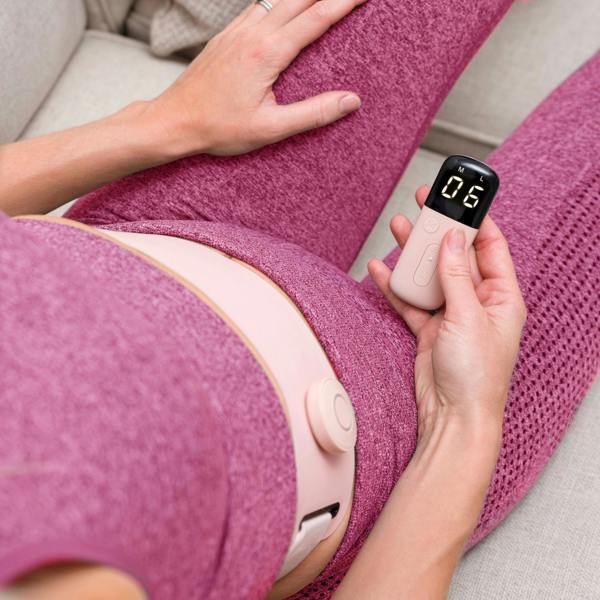 Wearing pink athletic wear, a person sits on a couch holding a small device displaying 6 and has the Ab, Tone & Shape EMS abdominal belt strapped around their lower abdomen for muscle stimulation.
