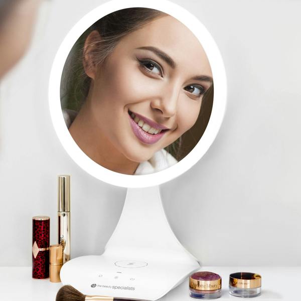 A woman smiles at her reflection in the Rio 5x magnification LED makeup mirror with wireless charging station on a white stand, surrounded by lipstick, mascara, a brush, and jars on a white surface.