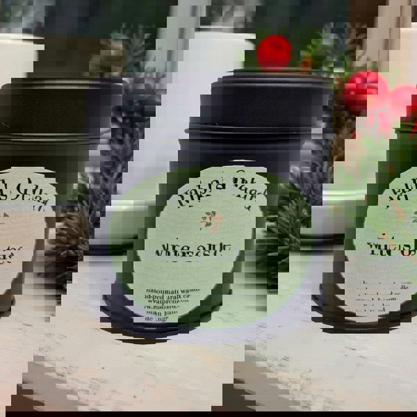 Candle in a black tin labeled 'Ralph's Orchard winter solstice' on a wooden surface with decorative elements.