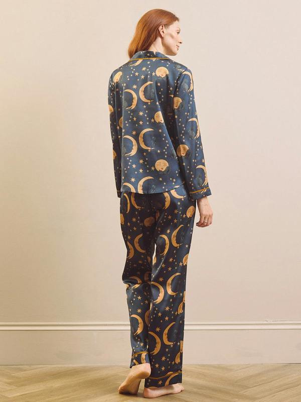 A woman stands barefoot indoors wearing a matching pyjama set with a dark blue background and an allover celestial print of gold crescent moons, stars, and planets, shown from the back.