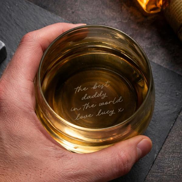 A hand holding a weighted tumbler filled with an amber-coloured spirit, looking down through the glass to reveal the base-engraved message 'the best daddy in the world love lucy x'.
