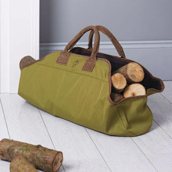 An open-ended green canvas log carrier bag packed with thick wooden logs, resting on a white wooden plank floor indoors.
