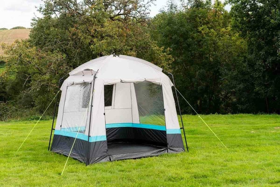 The OLPRO Pod Kitchen/Storage Tent pitched on a campsite with its door open and a tree line in the background.