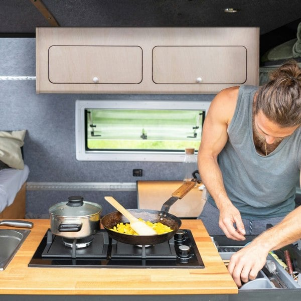 Overhead camper cabinet in light wood, featuring two upper storage compartments, above a compact kitchen with cooking utensils and food preparation