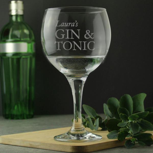 A personalised stemmed gin balloon glass by Abode Home & Gifts, featuring the custom script engraving "Laura's GIN & TONIC", filled with a clear drink and standing on a wooden board next to a green bottle and leafy eucalyptus sprigs.