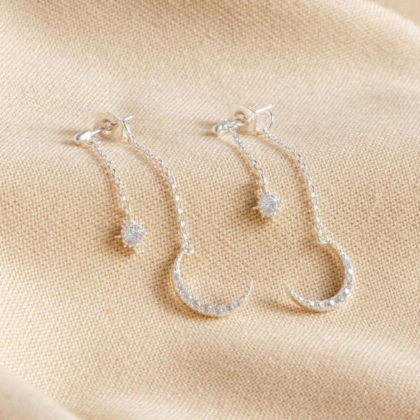 A close-up photograph of a pair of elegant silver-toned double drop earrings, featuring a smaller sparkling crystal star stud connected to a dangling larger crescent moon, set against a blurred, neutral background.