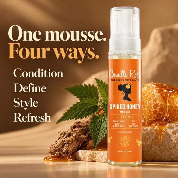 A bottle of Camille Rose Spiked Honey Mousse hair product stands on a warm beige surface with honeycomb, a honey dipper, and green leaves in the background. Large text on the left reads, 'One mousse.