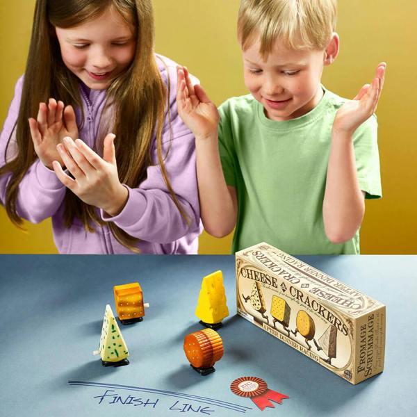 Two children standing at a table, cheering for wind-up toy racers shaped like cheese and crackers, with a "Finish Line" drawn on the table surface.