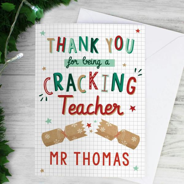 A white grid-patterned Personalised Cracking Teacher Christmas Card reading Mr Thomas at the bottom, resting on a white envelope next to holly leaves and festive fairy lights.
