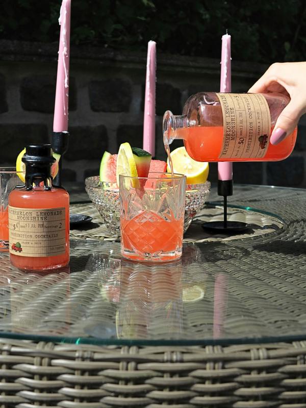 A hand pours a pink liquid from a glass bottle labeled 'Watermelon Lemonade Moonshine' into a clear glass tumbler on an outdoor patio table. Another bottle of the same pink liquid sits to the left, with pink taper candles and fruit garnishes in the background.