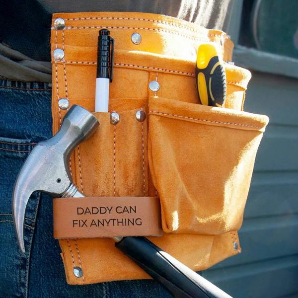 A close-up shot of an 11-pocket suede leather tool belt in a warm tan colour, worn over denim. The leather loop is clearly visible and engraved with the text "DADDY CAN FIX ANYTHING," with a hammer, pen, and screwdriver tucked into the pockets.