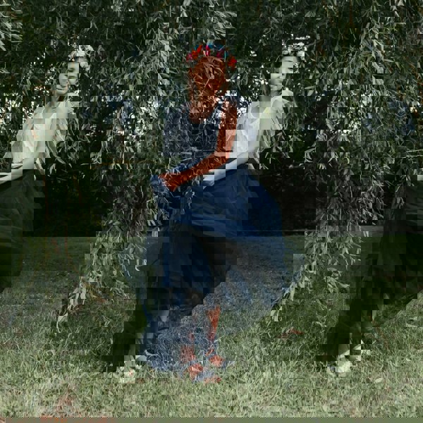 Young girl in a white top and navy blue skirt with a floral crown standing in front of green foliage.