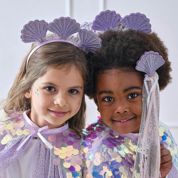 Close-up portraits of two young girls smiling while wearing matching purple shell mermaid headbands and tulle capes.