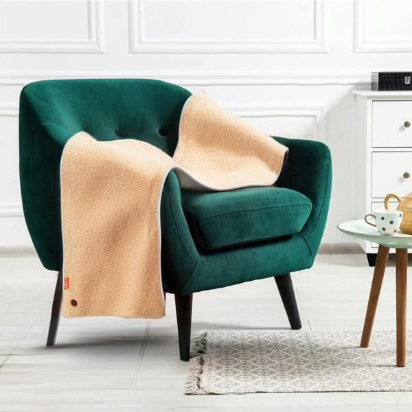 The COZY Heated Throw & Seat Cover Pad - JORV in beige is draped over a green velvet armchair beside a round table with tea and a cabinet, all set in a bright, modern room with white walls and a gray rug.