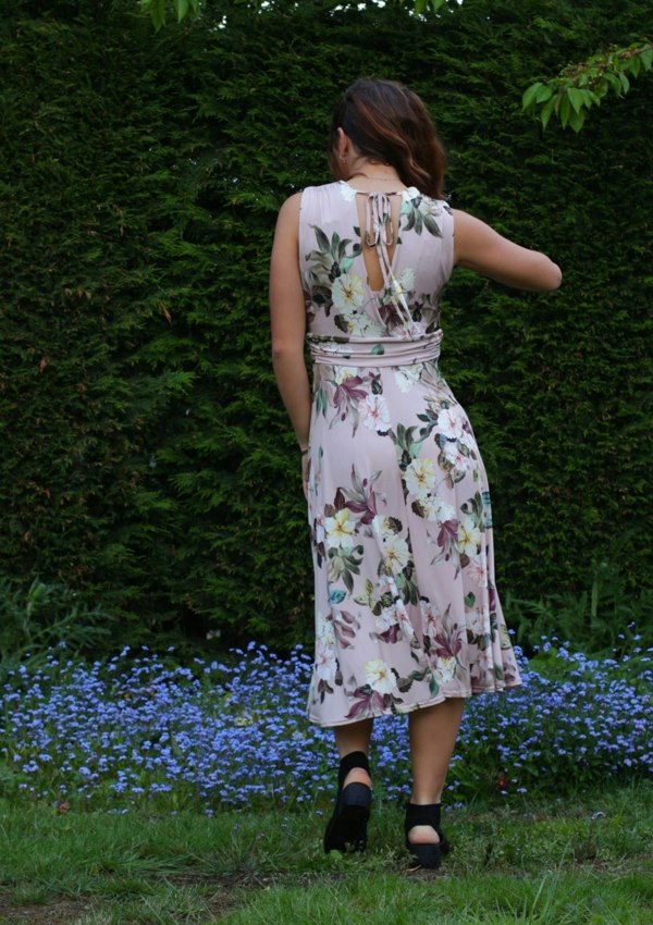 This is a young lady wearing a floral printed dress in pale pink. It has a mock V neck style top, which has gathers under the bust and round the back and the shoulders with an open back with a tie, secured into a bow. The model is wearing black lace up heels, she is facing away from us and she has her hand in her hair, silhouetted against a lush garden.