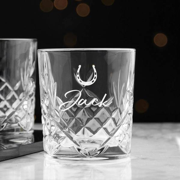 A diamond-cut crystal whisky tumbler engraved with a horseshoe and Jack in script font on a marble countertop.
