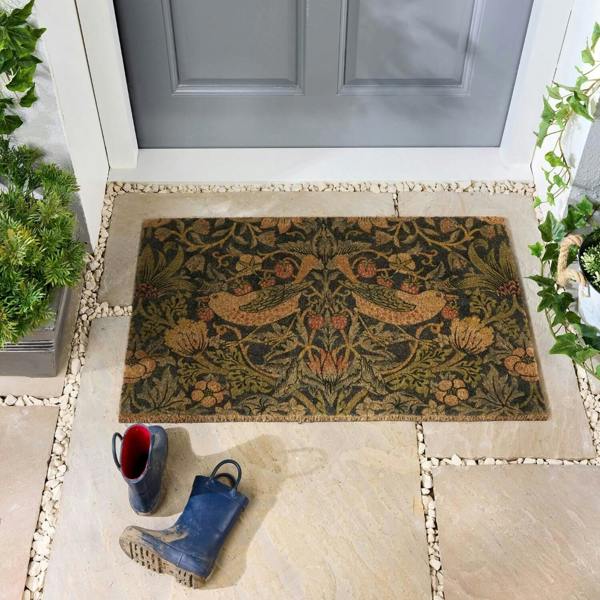 Decorative doormat with floral pattern in front of a gray door, with boots and plants on either side.