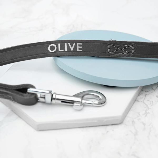 A personalised black leather dog lead stamped with the name OLIVE in silver foil next to a high-shine silver trigger clip.
