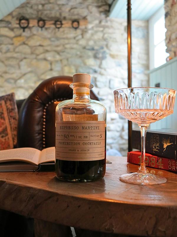 A bottle of Espreso Martini mix cocktail syrup with a cream-colored label, displayed on a rustic wooden table next to a cut crystal coupe glass in a cozy, warm-toned interior setting.
