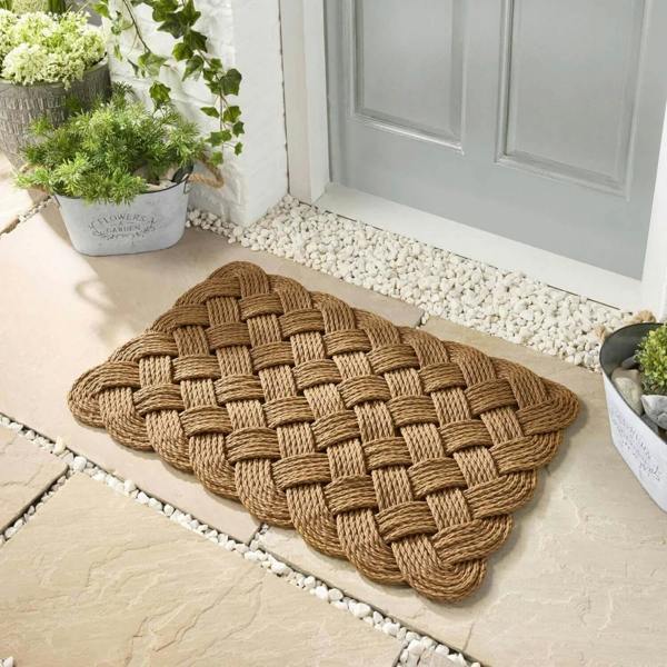 Woven doormat on a stone pathway with plants and a door in the background