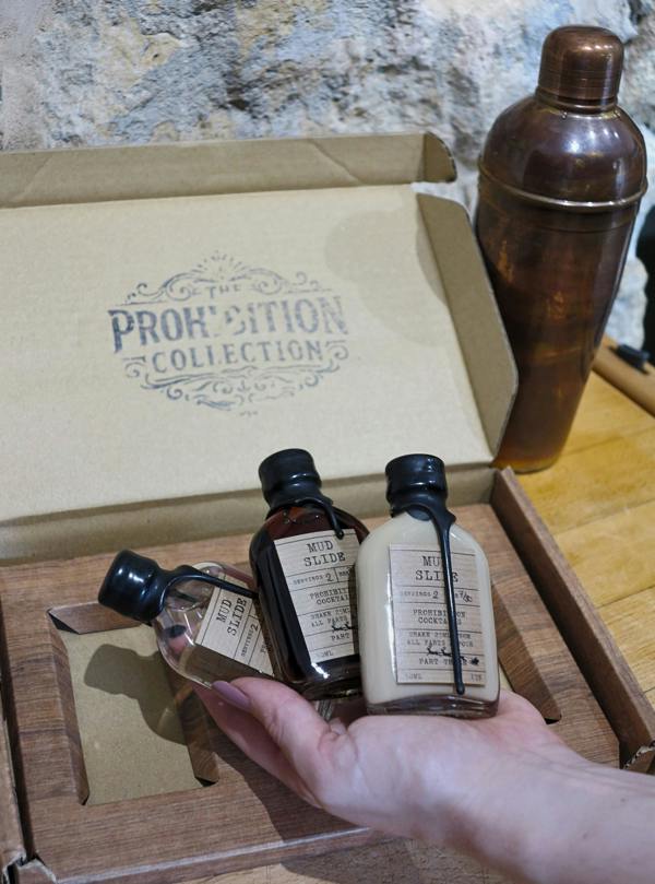 A hand holds three small bottles from The Prohibition Collection inside a wooden box, with a copper-colored cocktail shaker in the background against a stone wall.